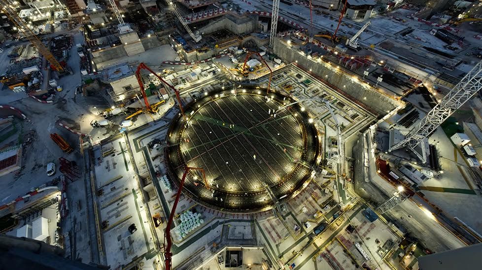 Aerial night view of a nuclear reactor foundation under construction, lit by floodlights, with red concrete pump booms reaching toward the circular reinforced base.